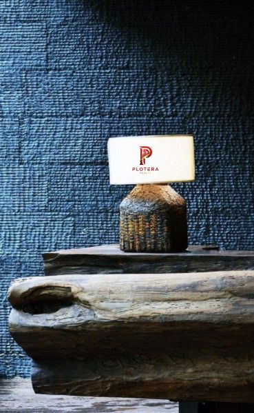 A moody, high-end interior vignette featuring a textured wallpaper in deep navy blue with a rugged, stone-like horizontal grain. The scene is lit by a small, artisanal table lamp with a rustic base, casting a warm glow against the dark wall, while a large, natural wood log sits in the foreground to emphasize the organic, raw-luxury aesthetic.